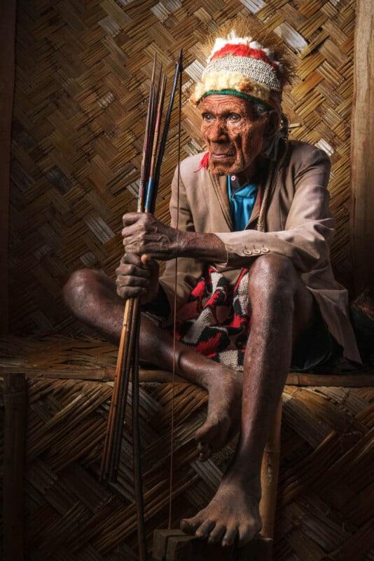 An elderly person sits barefoot on a woven mat in traditional attire, holding a set of arrows. They wear a colorful headdress and a blazer, with a determined expression, set against a rustic, woven backdrop.