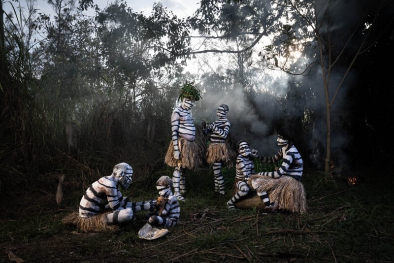 A group of people, painted in black and white stripes resembling zebra patterns, are gathered in a forested area. They wear grass skirts, with some sitting and others standing. Smoke can be seen in the background, partially obscuring the trees.