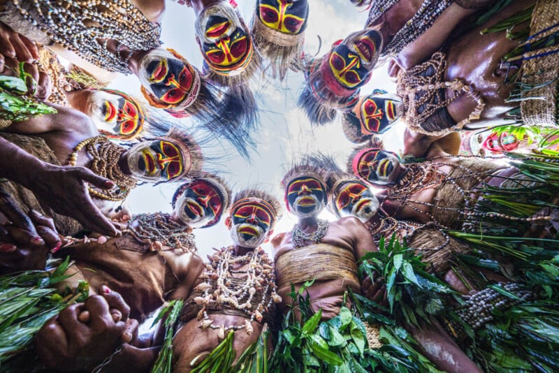 A group of people in traditional attire with brightly painted faces and headdresses form a circle, looking down at the camera. They wear costumes decorated with beads and leaves, set against a blue sky.