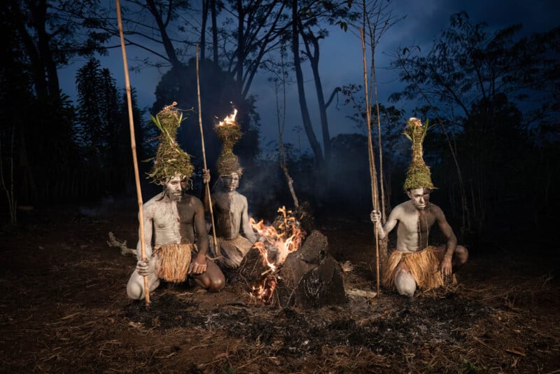 Three people dressed in traditional attire with body paint and grass skirts sit around a fire. They hold long sticks with leafy, flame-topped bundles. The background is a dark forest with silhouettes of trees against a dusky sky.