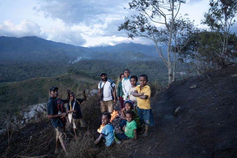 A group of people, including children and adults, gather on a hillside at dusk. They appear to be near a small fire with mountainous terrain visible in the background. Trees and a hazy sky complete the scene.