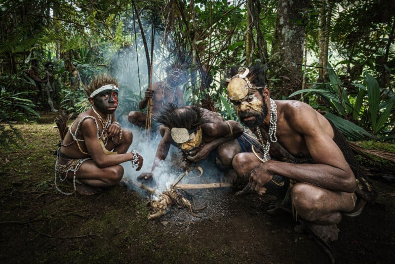 Four men in traditional attire, adorned with body paint and accessories, crouch around a small fire in a lush forest. Smoke rises as they perform a ritual or ceremony. Dense greenery surrounds them.