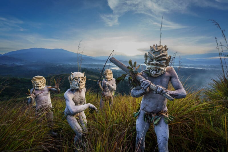 Four individuals wearing traditional tribal masks and body paint stand in tall grass against a backdrop of rolling hills and a cloudy sky. The scene conveys a cultural celebration or ritual in a natural setting.