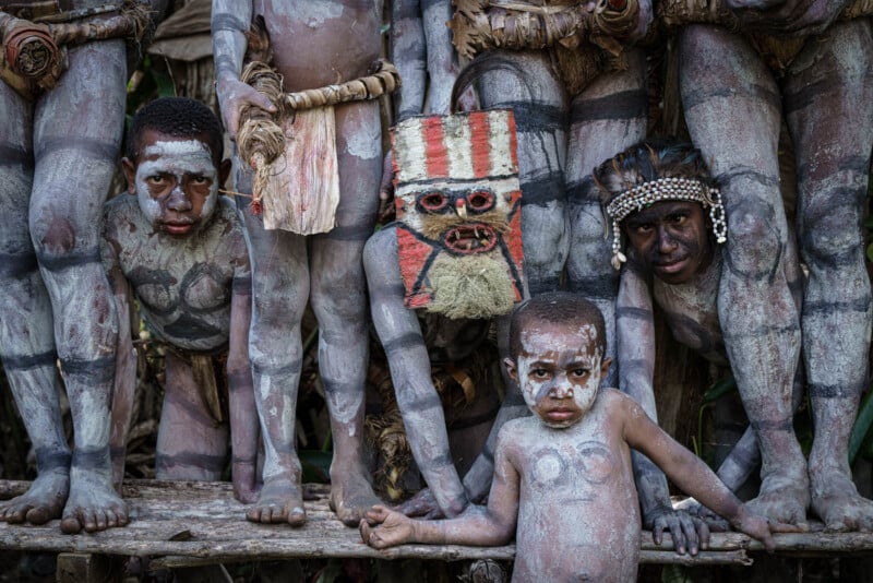 A group of people with body paint and traditional attire stand closely together. One person wears a mask with red stripes. A child in the foreground looks toward the camera. The setting appears natural with greenery in the background.