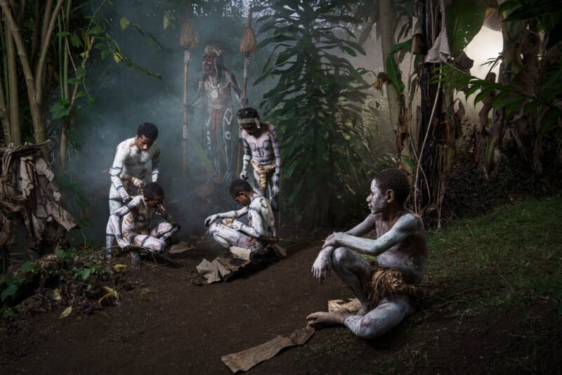 A group of people sits and stands in a lush, forested area with large leaves and mist. They are adorned with white body paint and traditional clothing. Some are crouched in discussion, and one sits alone on the ground, deep in thought.