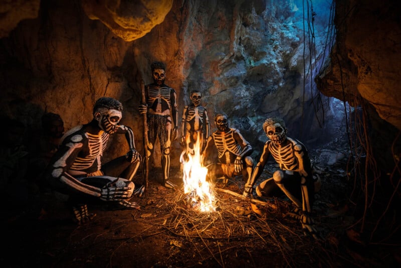 Five individuals in a cave are painted with skeleton designs and sit around a small fire. The cave is dimly lit, with shadows and a bluish glow in the background, creating an atmospheric contrast between light and dark.