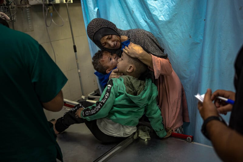 A woman embraces two distressed children in a medical setting. One child is seated on a stretcher wearing a green jacket, while the other child leans on the woman. Medical professionals are nearby against a blue privacy curtain.