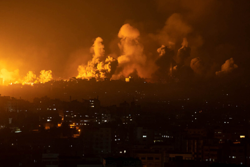 A city skyline at night is illuminated by multiple fireballs and explosions. Thick smoke rises against a dark sky, suggesting intense conflict or bombardment. City lights are visible below the scene, contrasting with the fiery destruction above.