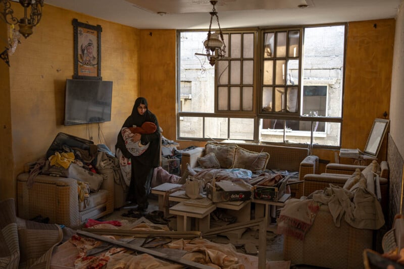 A woman in a black robe holds a child, standing in a room with damaged furniture, scattered debris, and broken windows. The walls are yellow, and a poster hangs crookedly. The atmosphere is somber and chaotic.