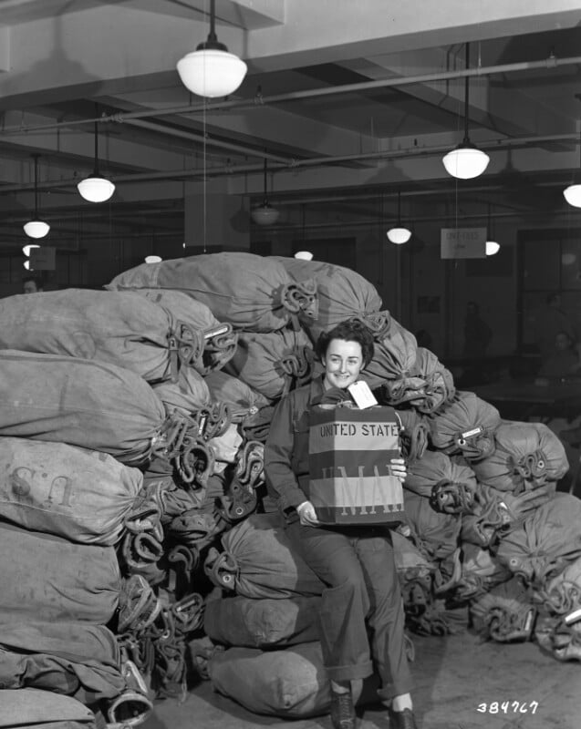 A person smiling and holding a large sack labeled "United States Mail" in a room filled with piles of mail bags. The room is lit by several ceiling lights, and there are signs indicating other sections in the background.