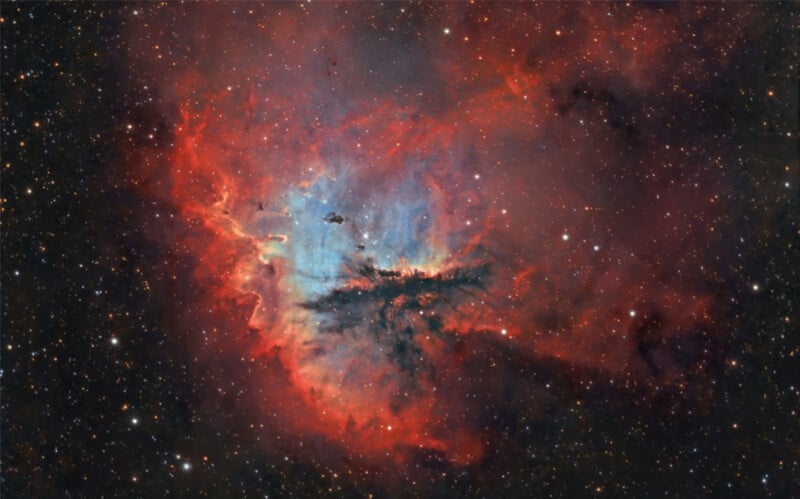 A vivid image of the Pacman Nebula, featuring bright red and blue clouds of interstellar gas and dust. The center is illuminated, resembling a partially opened mouth, surrounded by a field of stars against the dark backdrop of space.