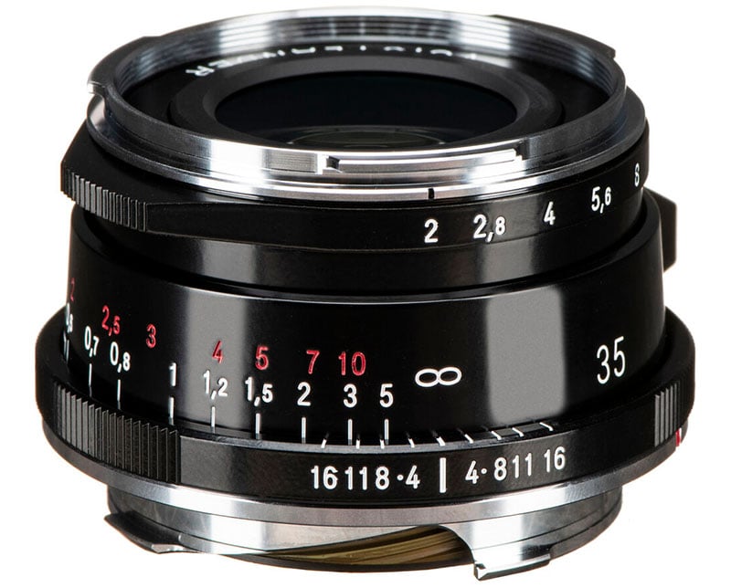 Close-up of a black camera lens with aperture and focal length markings in white and red. The lens has a shiny metal mount and manual focus ring, displaying numbers and symbols for different settings.