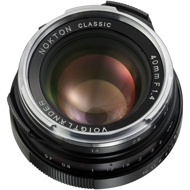 A close-up of a Voigtländer Nokton Classic 40mm f/1.4 camera lens. The lens barrel is black with silver accents, featuring engraved focus and aperture scales. The glass element reflects light, showing a slight color gradient.