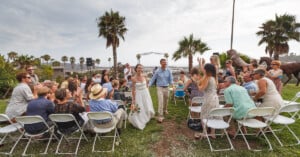 A bride and groom walk down the aisle outdoors while guests cheer. They're surrounded by palm trees, with an arch adorned with flowers in the background. Guests are seated on white folding chairs, and the setting is grassy and tropical.