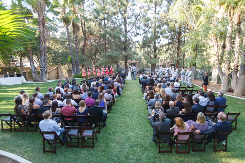 Outdoor wedding ceremony with guests seated in rows on a lawn. A couple stands at a wooden arch surrounded by trees. Bridesmaids in pink dresses are on the left, and groomsmen in light suits are on the right.