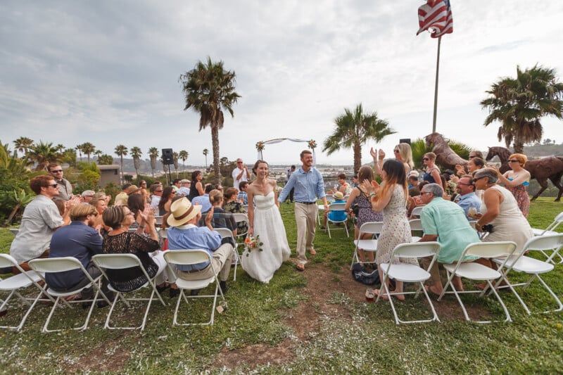 A bride and groom walk down an aisle outdoors, smiling at guests seated on either side. Palm trees, an American flag, and a scenic view are in the background. The setting suggests a relaxed, joyful wedding ceremony.