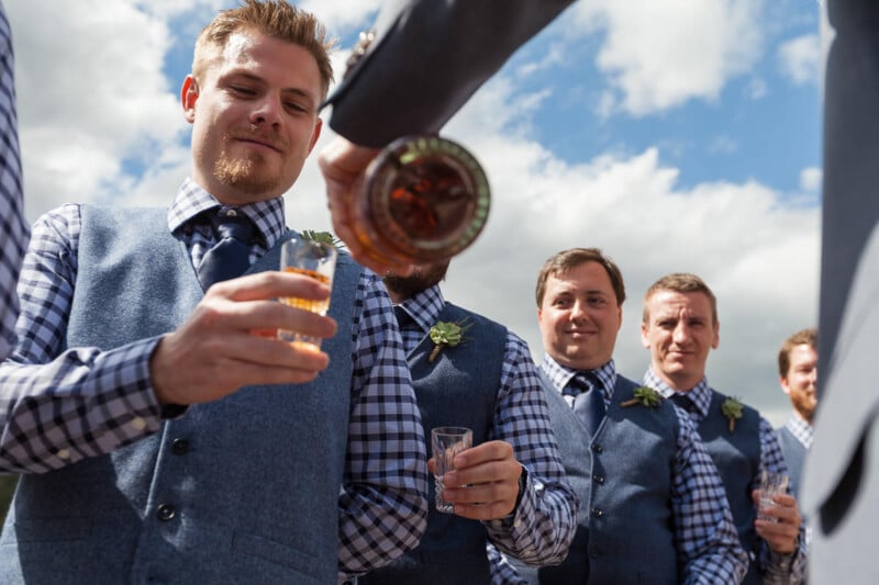 A group of men in checkered shirts and blue vests, standing outside under a cloudy sky, receive drinks from a bottle being poured. Some wear boutonnieres as they smile and prepare for a toast.