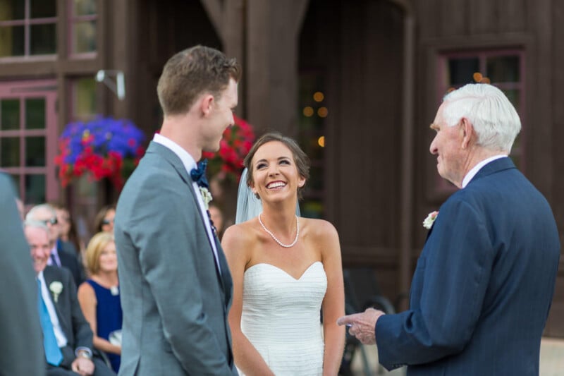 A bride in a strapless white dress smiles at her groom, who is wearing a gray suit and bow tie, during an outdoor wedding ceremony. An elderly man in a dark suit stands before them, speaking. Guests are seated in the background near colorful flowers.