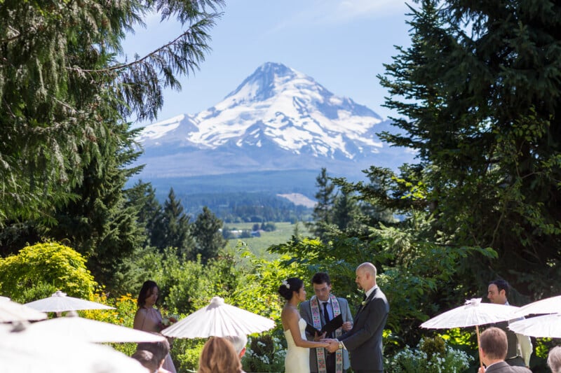 A couple stands facing each other during an outdoor wedding ceremony with a snow-capped mountain in the background. Guests are seated under white umbrellas, surrounded by lush greenery and trees.