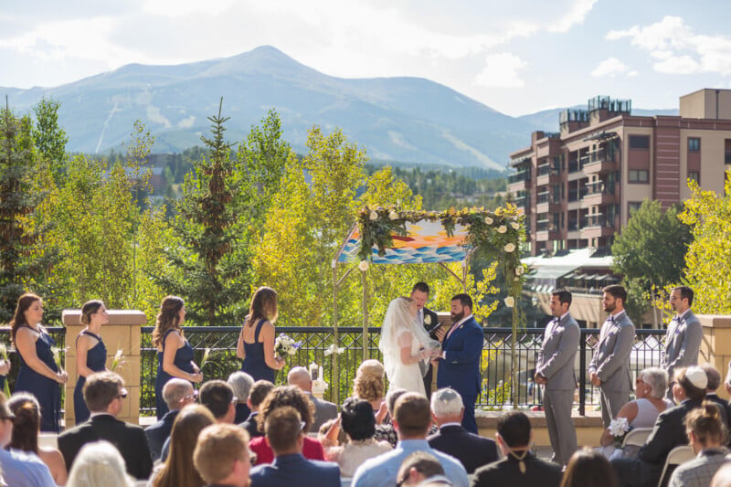 An outdoor wedding ceremony with a couple exchanging vows under a floral arch. Guests are seated, and the bridal party stands on either side. In the background, there's a scenic view of mountains and trees under a clear, sunny sky.