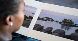 A person is looking at an open photo book displaying serene landscape images of a rocky shoreline and calm waters under a pastel sky. A small island with trees is visible in the distance.