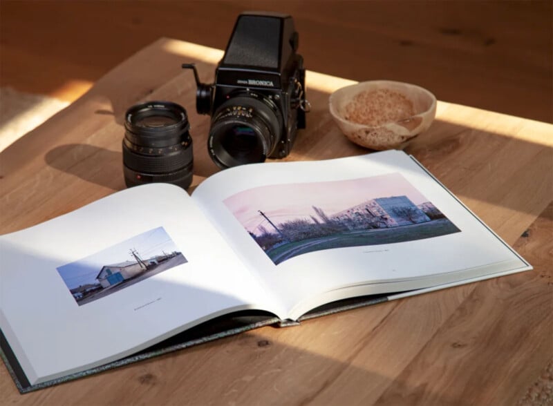 A camera and two lenses rest on a wooden table next to an open photography book with landscape photos. A small bowl with a wooden spoon is nearby, and sunlight casts a soft glow over the scene.