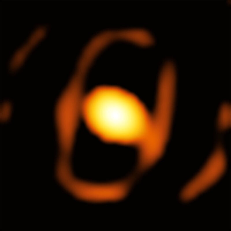 A glowing orange oval shape with abstract, irregular patterns against a black background, resembling a blurred, artistic depiction of a nebula or celestial object in deep space.