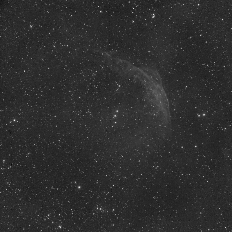 A black and white image of a starry night sky featuring a faint, wispy nebula among numerous scattered stars. The nebula appears as a subtle, curved structure in the upper right area of the image.