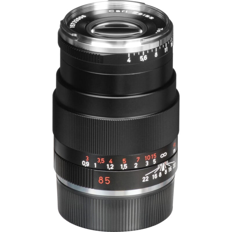 A black camera lens with silver and red detailing, featuring various focus and aperture measurements. The lens has textured grip sections and an engraved brand name on the front rim.