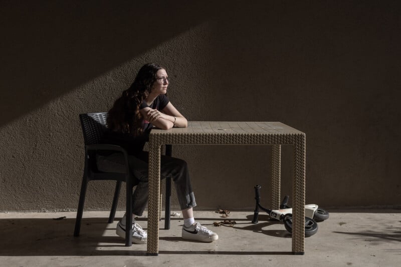 A person with long, dark hair sits at a woven table in a chair, looking thoughtfully to the side. They are in a shadowed area with a scooter lying on the ground nearby. The scene is lit by a soft beam of sunlight.