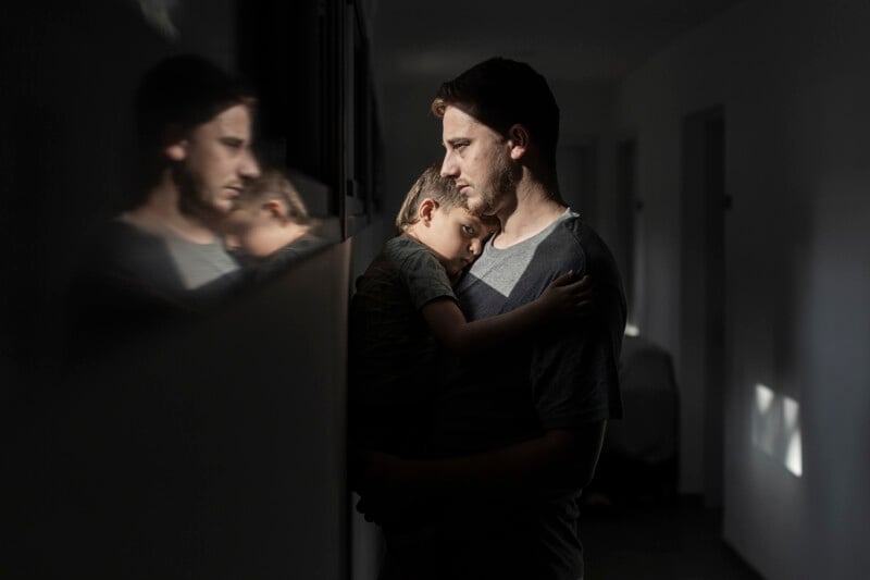 A man holds a young boy close in a dimly lit hallway, both looking thoughtful. Their reflection is visible on a glass surface, adding depth to the scene. Light filters in from the side, highlighting the tender moment.