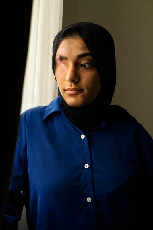 A person wearing a blue shirt and a black hijab looks thoughtfully out of a window with light streaming in. The room appears dimly lit.