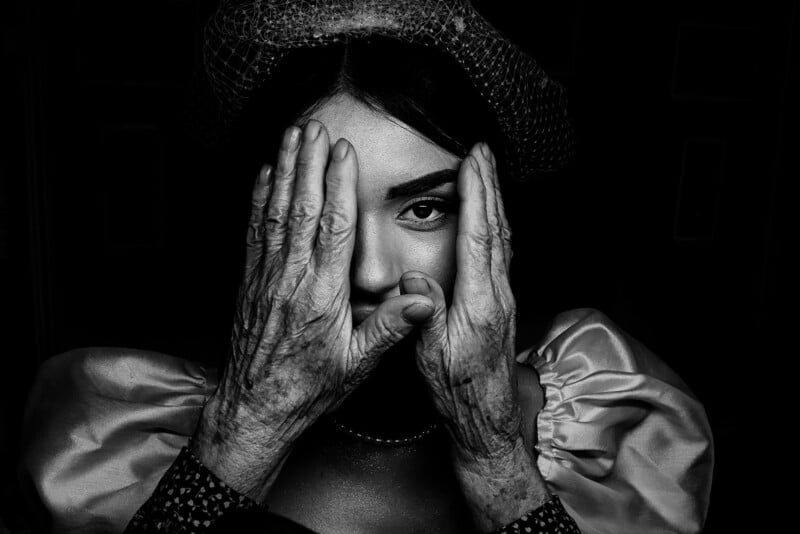 A woman in a vintage hat and dress looks directly at the camera. Her face is framed by two elderly hands, creating a dramatic contrast between youthful and aged features in black and white.