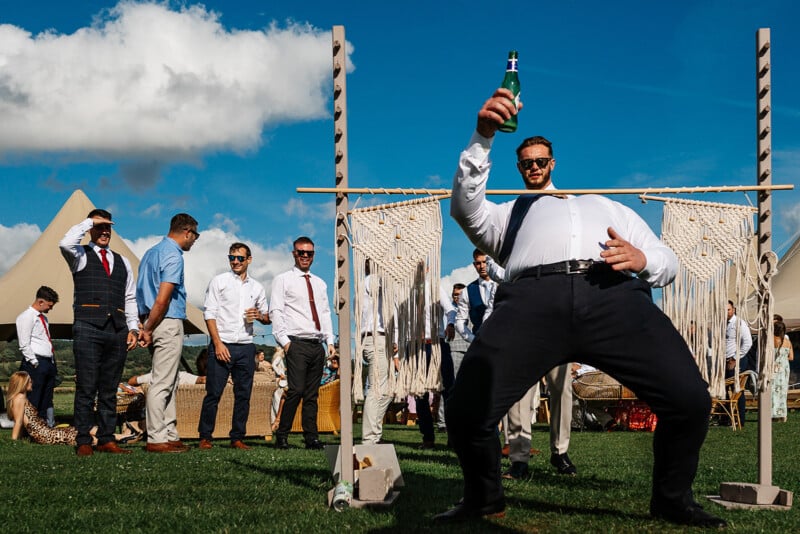 A group of men outdoors at an event with a tent in the background. One man in the foreground is limbo dancing under a macrame bar while holding a beer bottle. The sky is blue with a few clouds.