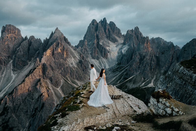 A couple in wedding attire stands on a rocky mountain ridge with jagged peaks in the background. The sky is overcast, adding dramatic lighting to the scene. The bride's dress flows gracefully behind her.