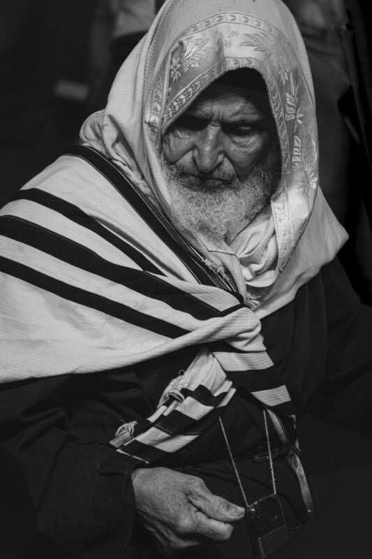 A⁤ black and white image of an elderly person with a long beard,⁣ wearing a ‌headscarf and traditional‌ clothing, possibly Middle Eastern. They‌ look down thoughtfully, holding a stringed object.