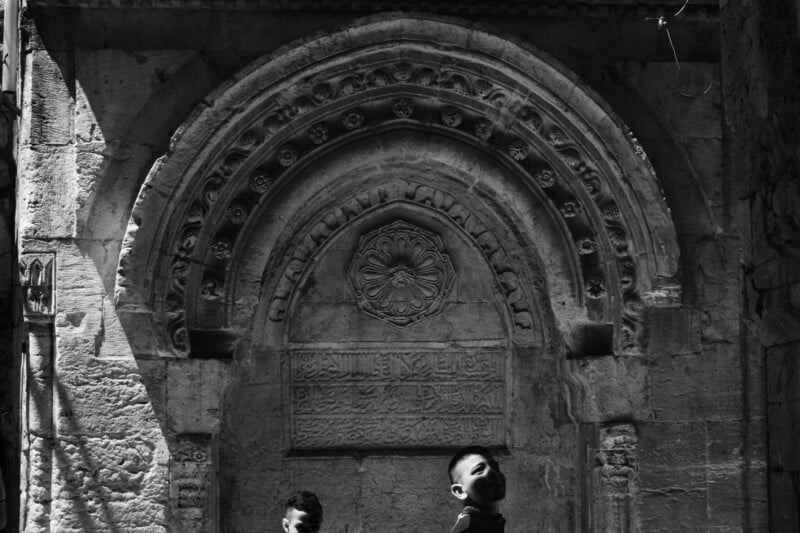 Two children stand in front of an ancient stone archway adorned with intricate carvings and inscriptions. The sunlight casts dramatic shadows, highlighting the ornate details of the architecture. The image is in black and white.