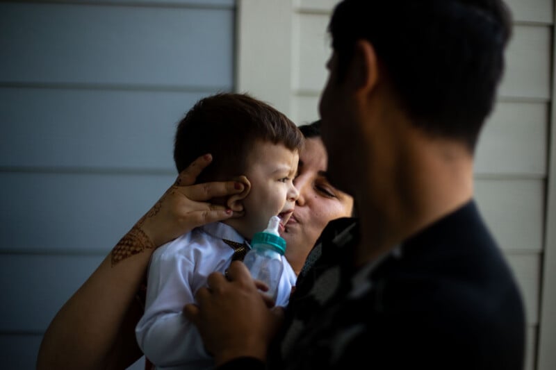 A woman holds a toddler in a white shirt close to her face, gently cradling him as he drinks from a baby bottle. Another person is partially visible, standing nearby. The setting appears to be indoors.