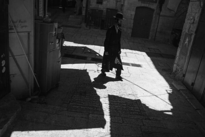 Black and white ⁢image of a person walking on a cobblestone street in a shadowed⁢ alley. The individual wears a hat and⁣ long ⁢garment while⁢ holding an umbrella. Sunlight and shadows create contrasting ⁢patterns on the ground.