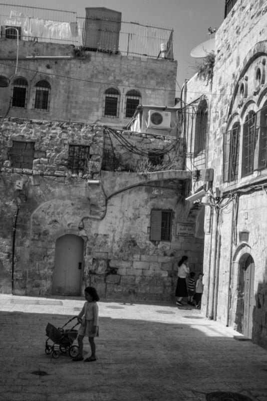 A black and white scene of an old stone courtyard with a small child pushing a stroller in the foreground. In the background, a woman stands near a door with two children. The buildings have arched windows and visible wiring.