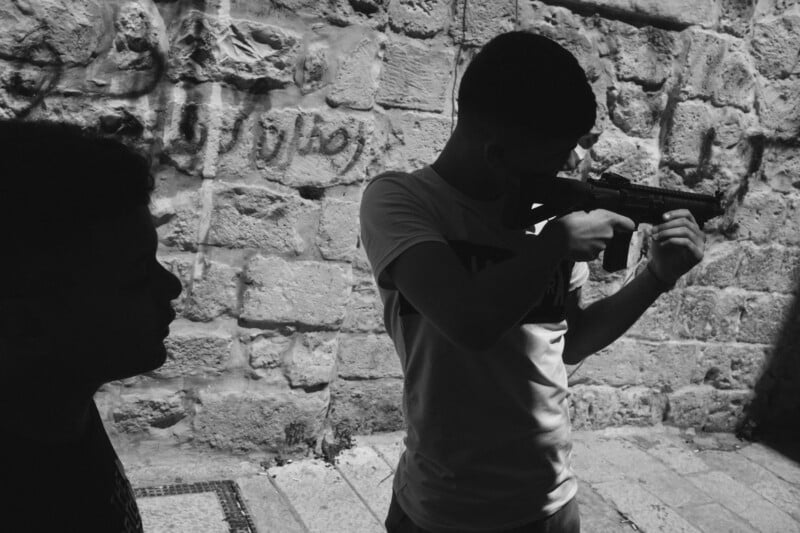 Black and white photo of two boys near ‌a ​stone wall. One ⁢boy, in the foreground, holds a toy gun‌ and poses as if aiming. The other boy stands in silhouette, ‍watching the first boy. Graffiti is visible on ‍the wall.