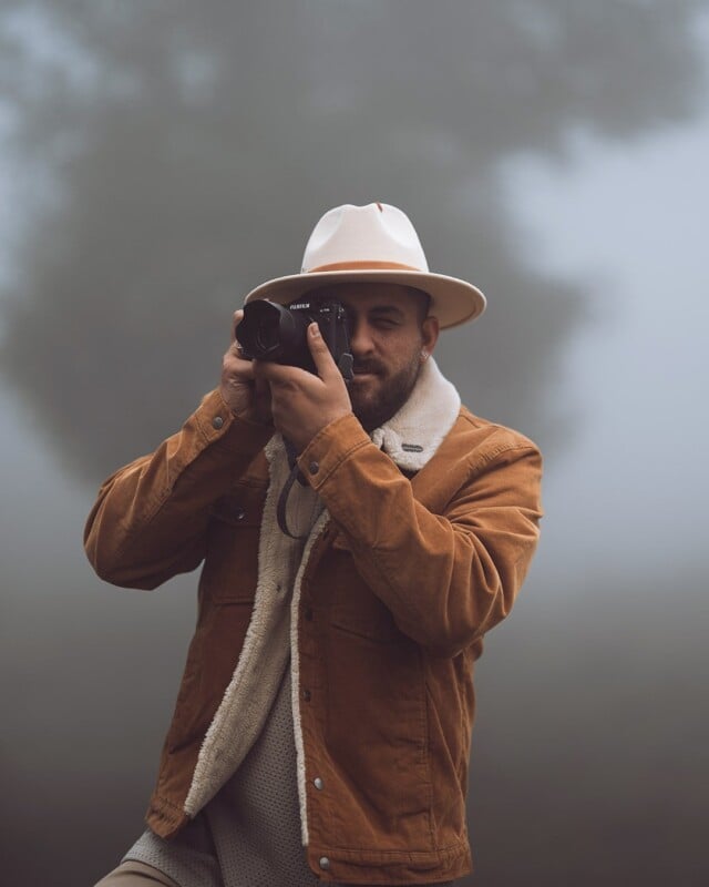 A person wearing a brown jacket and white hat is using a camera. They are standing outdoors on a foggy day, with blurred trees in the background.