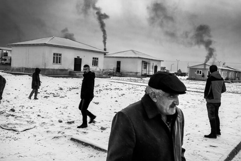 A black and white photo shows several people walking on a snowy ground near small houses with smoke rising from chimneys. The sky is cloudy, and the scene conveys a cold, wintry atmosphere.