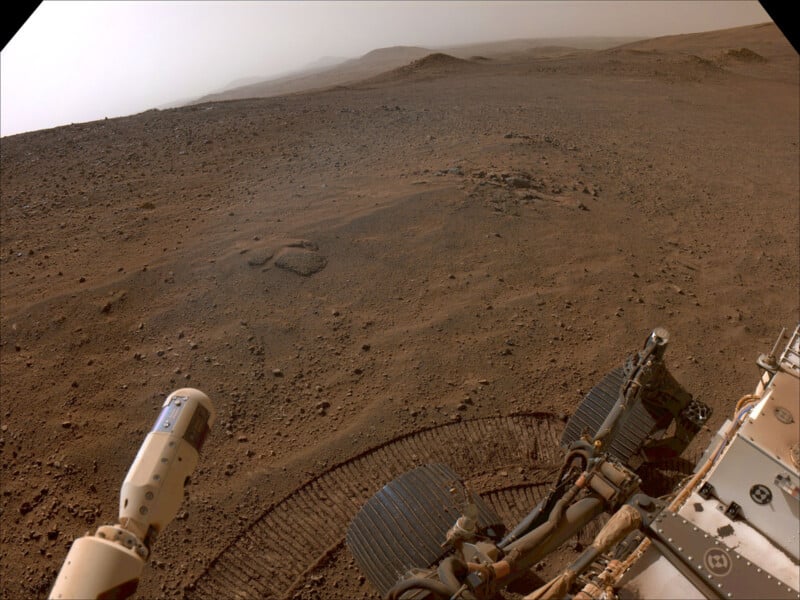 A dusty Martian landscape with rocky terrain and hills in the background. In the foreground, part of a rover with visible wheels and instruments is capturing the view. The sky appears hazy, suggesting a dusty atmosphere.