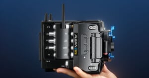 A hand holds a professional Arri Alexa 65 camera against a dark blue background. The camera features various control dials, buttons, and a blue lever, showcasing its advanced technology and sleek design.