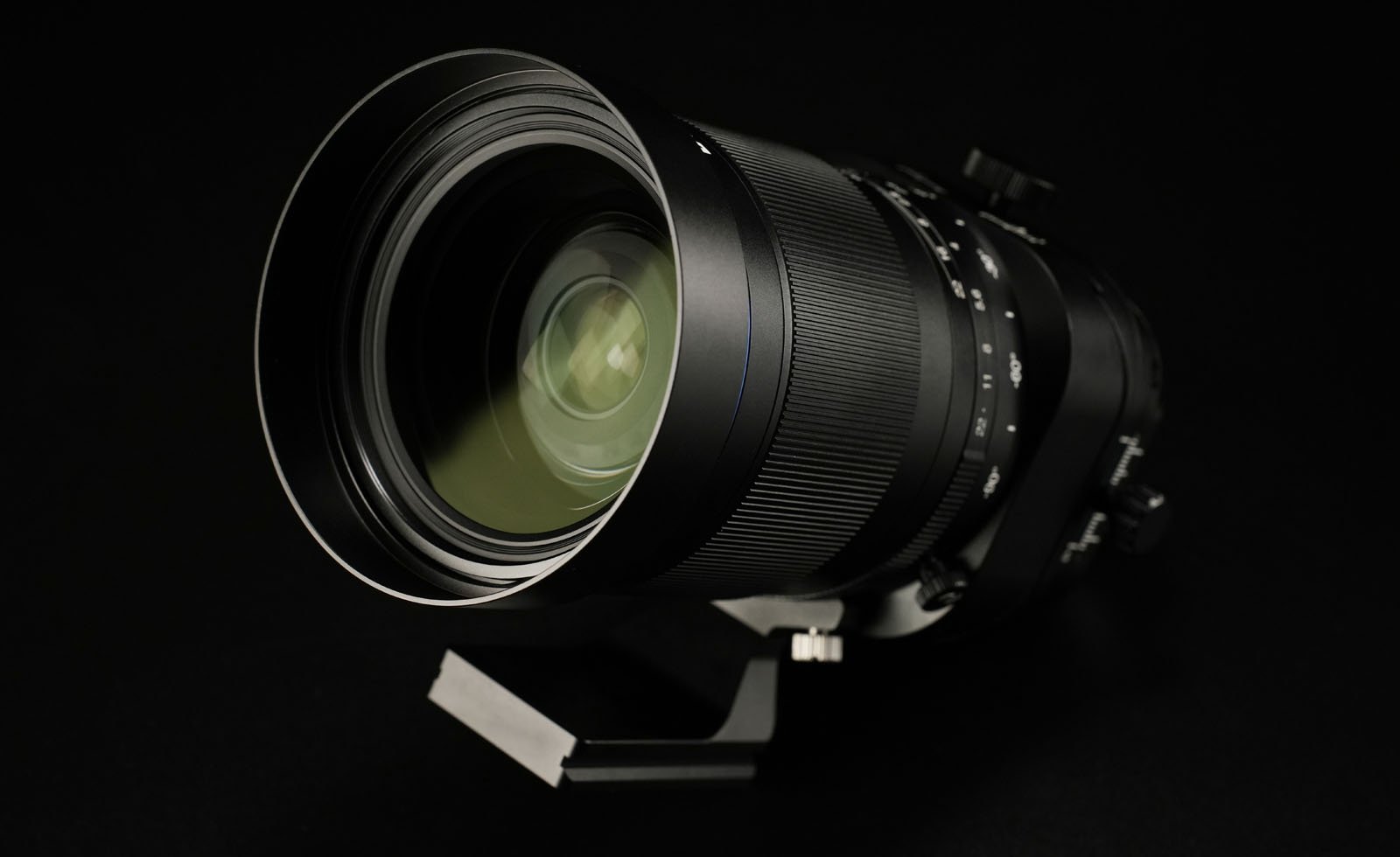 Close-up of a large black camera lens with visible details, including the focus ring and mounting bracket. The lens is angled slightly off-center against a dark background, highlighting its sleek design and smooth texture.