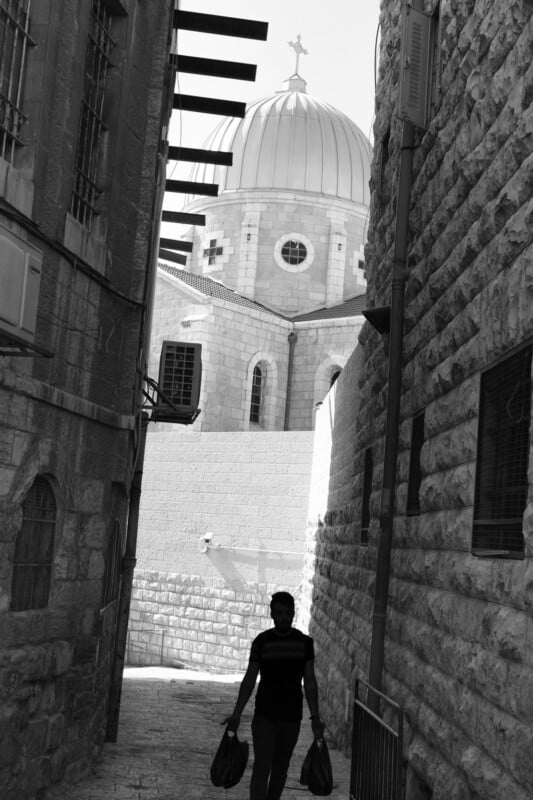 Black and white photo of a⁣ person carrying ​bags, walking ⁢through⁤ a narrow‍ alley between stone buildings, leading to ‍a large building with a dome and ⁣a cross on top.⁢ Sunlight casts shadows across the scene.