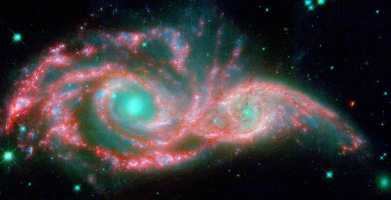 A vivid image of two interacting galaxies creating a swirling pattern. Bright teal cores are surrounded by pink and red spiral arms, set against a dark space filled with scattered stars.