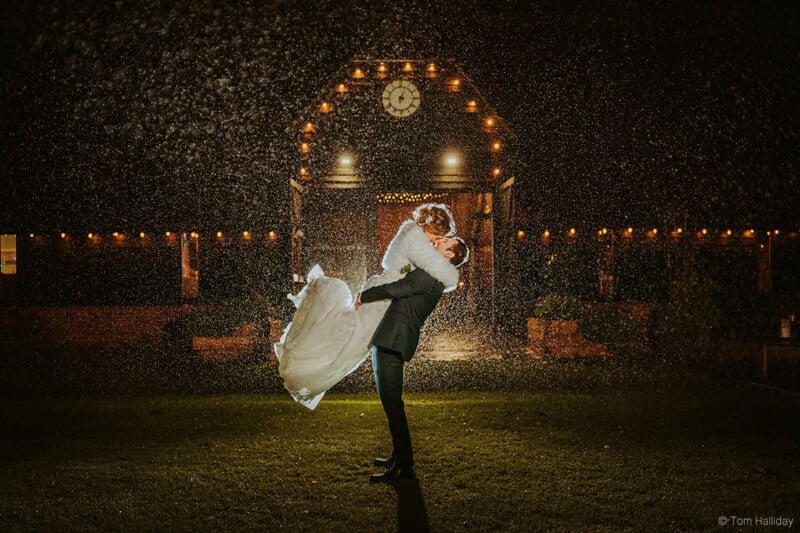 A couple in a wedding dress and suit embrace, with the groom lifting the bride, surrounded by glowing lights and falling snowflakes against a dark sky. A building with a clock is illuminated in the background.