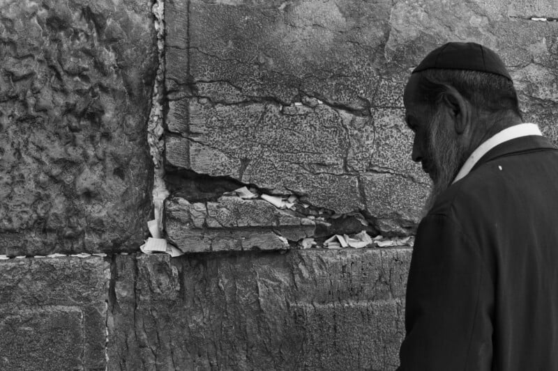 A man in ​a traditional skullcap stands in ​profile facing the⁣ Western⁤ Wall, which is filled with notes in its crevices.​ The wall is made of large,textured stones. The scene is in black and ⁢white.